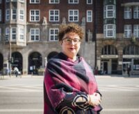 Eine Frau steht vor einem Backsteingebäude und schaut in die Kamera.